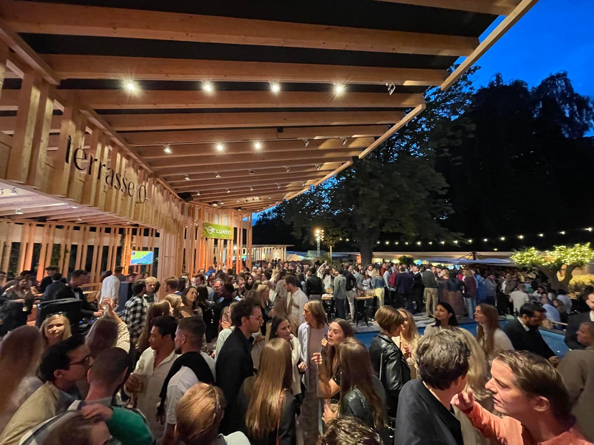 A full terrace of guests talking under a lit wooden pergola at dusk